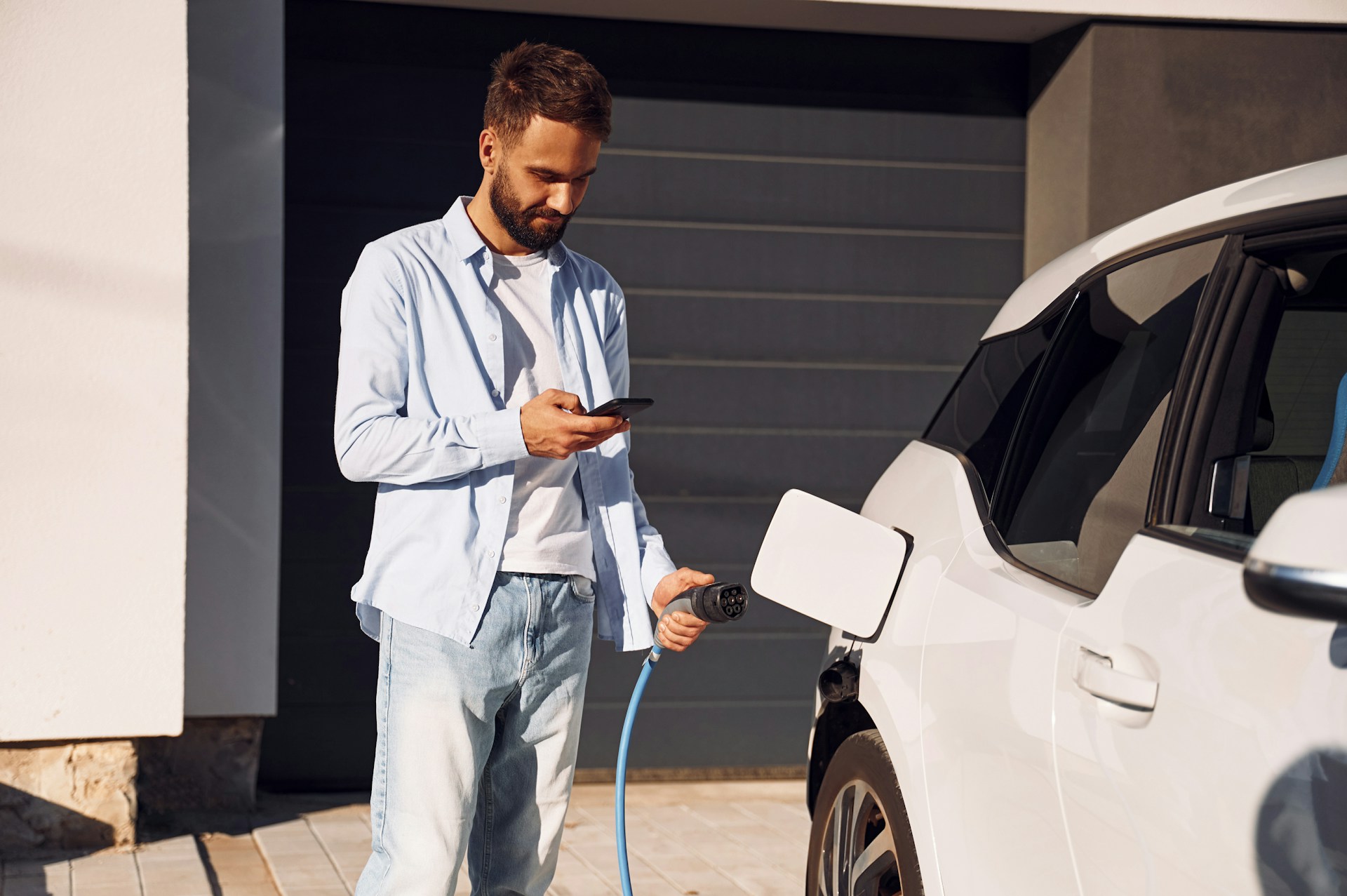 Man using smartphone while charging electric vehicle at home, highlighting the growing trend of EV charging station installations for homeowners.