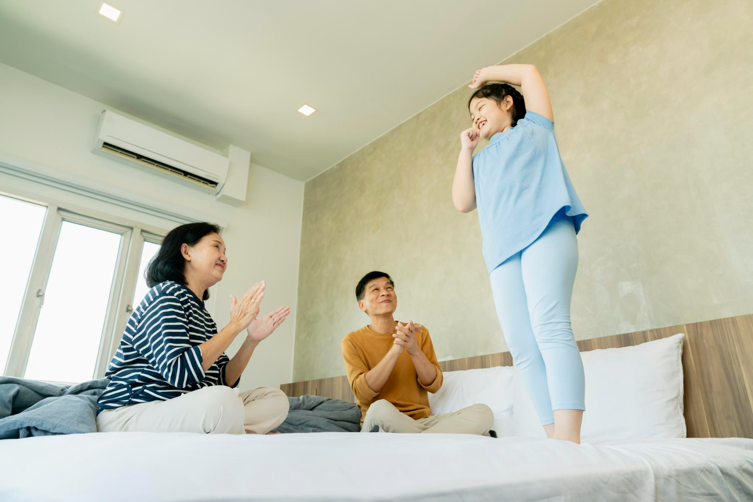 Family enjoying time together in a bedroom, with a child playfully standing on the bed, while parents applaud, showcasing a comfortable home environment.
